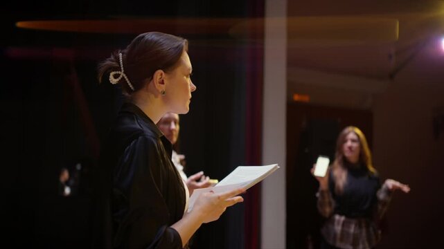 Young actors rehearsing dramatic scene on dimly lit theater stage, following script pages under warm spotlights during an intimate performance practice session
