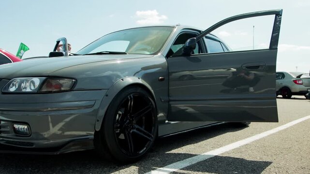 Close up view of modified dark grey sedan car parked outdoors on asphalt ground with white lines. Vehicle features wide body fender flares, black multi spoke alloy wheels. Car door open. Scene capture