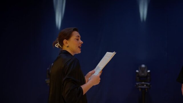 Young actors rehearsing dramatic script on dimly lit theater stage, holding pages and phones while preparing lines and performance under focused stage lighting
