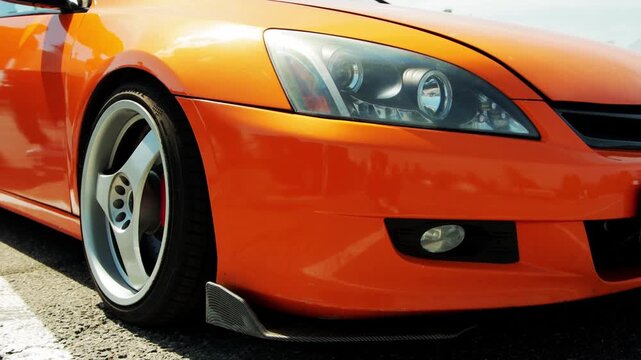 Orange modified sport coupe, front view, parked outdoors. Custom silver wheel, low-profile tire, clear headlight, fog light, carbon fiber front lip. Sunlight on glossy paint.