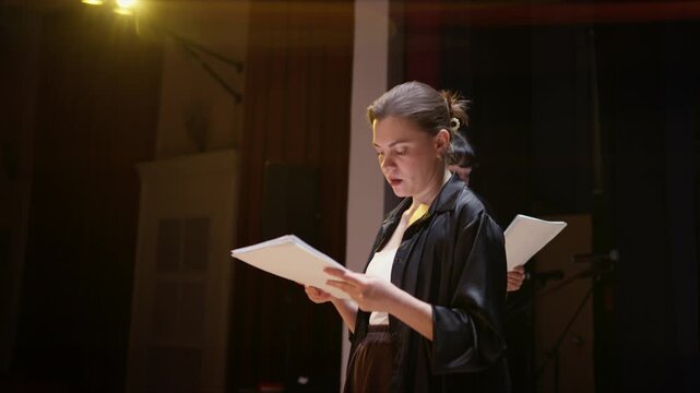 Focused performers stand on a dimly lit stage reading from scripts during a dramatic rehearsal, illuminated by warm spotlights in a quiet theater setting