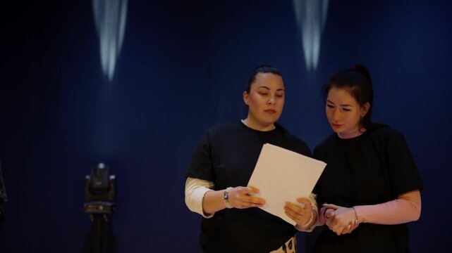 Focused performers rehearsing lines together on a dimly lit stage, collaborating with scripts in hand under dramatic theater lighting before a group performance