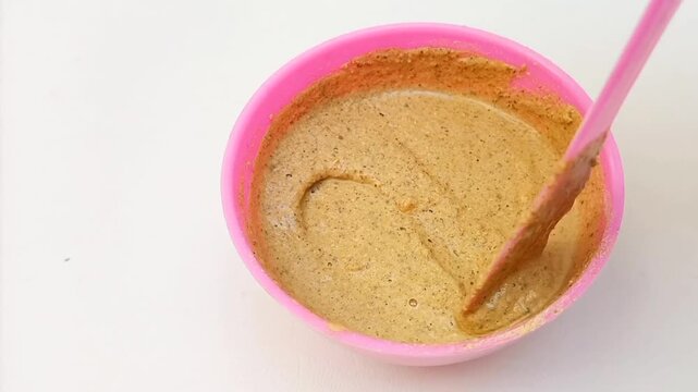 Traditional Ayurvedic Ubtan Powder Mixing Process in Pink Plastic Bowl with Mixing Spoon Isolated on White Background