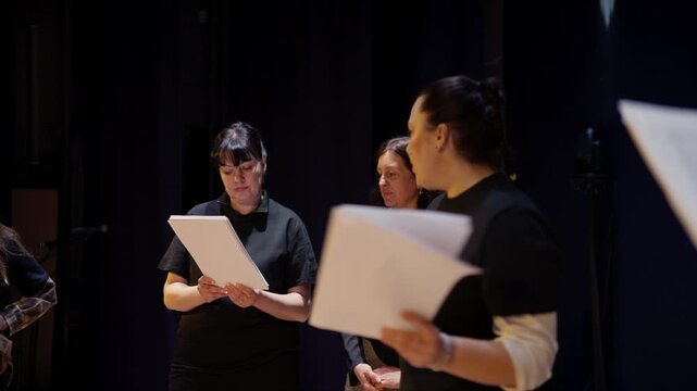 Group of adult performers rehearsing a script on a dimly lit stage, reading from printed pages and collaborating during a dramatic theater practice session