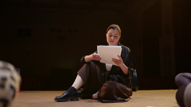Thoughtful woman seated on dimly lit stage reading from paper during intimate performance rehearsal, focused expression suggesting storytelling or dramatic monologue