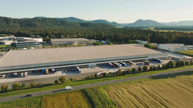 Large logistics center shows trucks loading and unloading cargo in the distribution area with mountains in the background and clear skies.