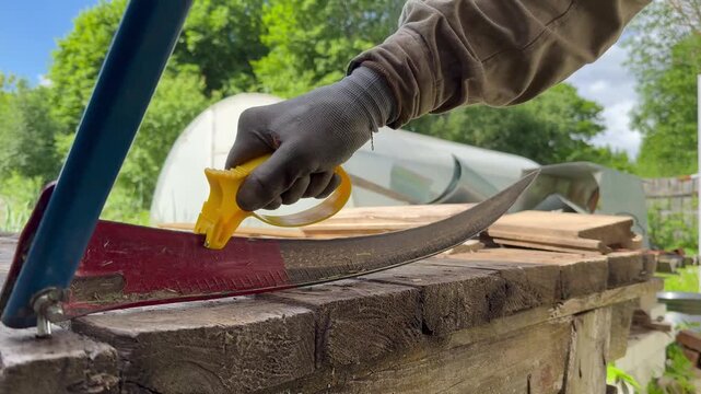 Close-up of a worker in protective gloves sharpening a manual scythe blade with a yellow sharpener on a wooden workbench. 