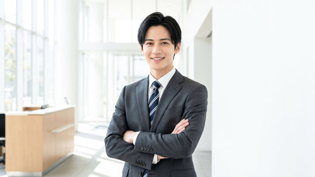 明るいエントランス空間で腕を組み、親しみやすい笑顔を見せる若いビジネスマンのポートレート。清潔感のあるスーツスタイルと開放的なオフィス背景が、信頼感、採用、営業、コンサルティング、企業案内、キャリア、ビジネスコミュニケーションのイメージ訴求に適した高品質な人物素材