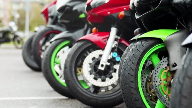 Close-up view of front wheels of multiple sport motorcycles parked in a line on asphalt road, highlighting brake discs and colorful rims. Scene depicts vehicle lineup during day with strong lines of m