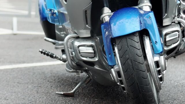 Close up view of motorcycle front section. Bright blue fender shows water droplets, indicating recent rain or wash. Silver engine components, chrome accents, black tire. Vehicle parked on asphalt grou