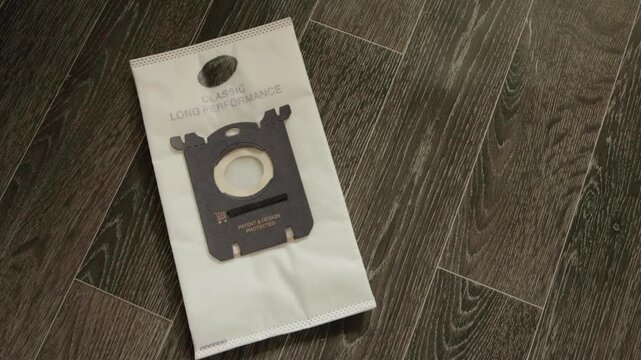 Close up view of person hand removing old vacuum cleaner dust bag filled with dirt. New empty dust bag prepared for installation on dark wood parquet floor. Essential chore.