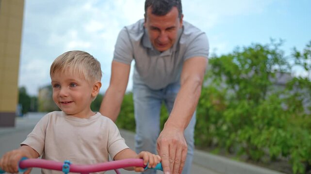 Father helps little son ride bike on street. Happy boy holds handlebars while dad pushes tricycle. Parent teaching kid to ride bike outdoors. Father and son enjoy active family time together