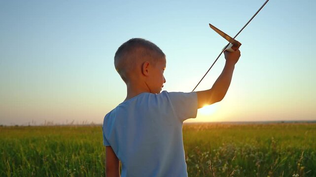 Little boy holds a toy airplane in a green field at sunset. Child plays with a wooden plane in nature. Kid dreams of becoming a pilot during summer evening. He launches the glider outdoors.
