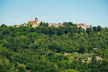 Fototapeta premium Mondaino village on its hill in Montefeltro, Italy, with the church of San Michele Arcangelo rising above green forest under clear sky