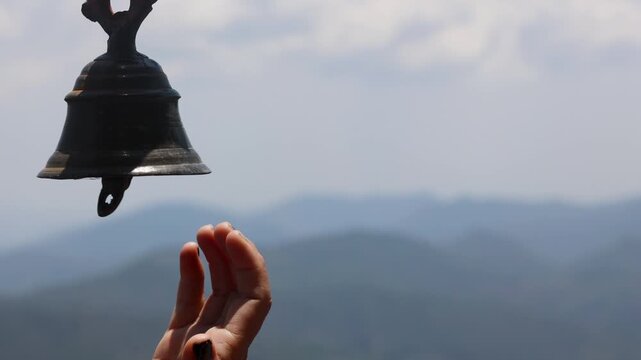 Raw video footage of a hand ringing a traditional temple bell with blurred mountain scenery in rural Uttarakhand, India. Spiritual atmosphere and peaceful countryside devotion.