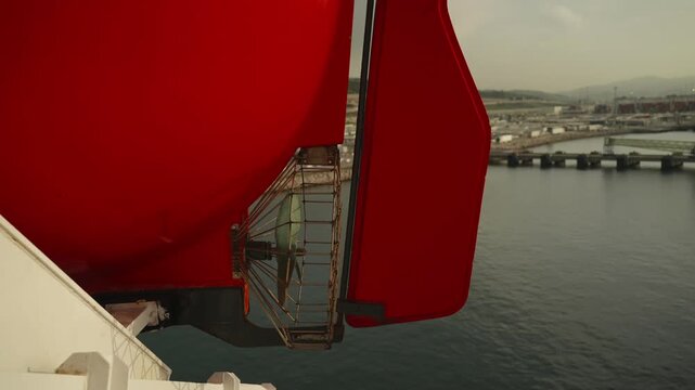 Red rescue lifeboat secured in davit cradle on ferry deck with industrial port and sea visible behind