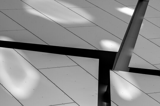 Black and white photograph studies ceiling panels, light reflections and diagonal beam, making interior architecture a graphic surface