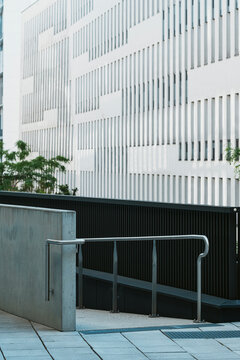 Metal handrail follows concrete ramp below white facade, with trees and urban architecture forming a clean exterior access detail