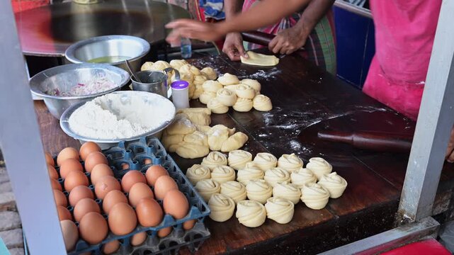 Local cooks roll paratha dough on a wooden counter next to fresh eggs and flour bowls at a Bangladesh food stand. Documents traditional street food preparation, market business, and daily trade.