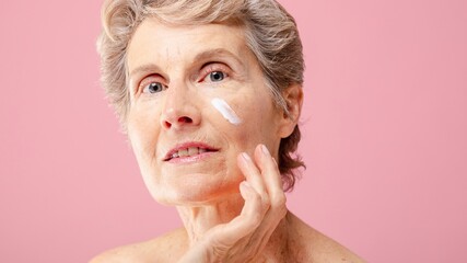 Senior white woman applying skincare cream on her face against pink background