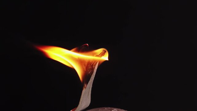 Macro view of paper burning slowly with bright orange flames in dark environment.