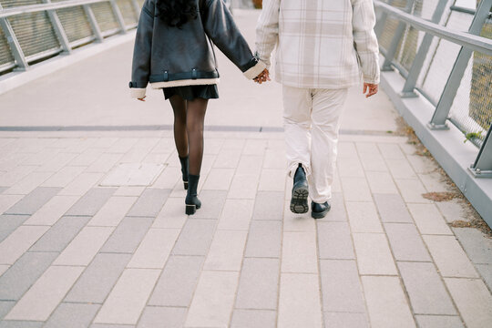 Individuals holding hands on urban bridge scene. Pair of lovers stroll hand in hand over tiled bridge amid overcast cityscape with casual attire