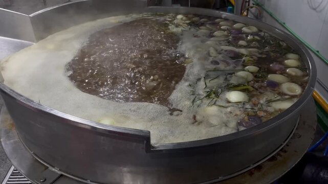 Stock broth boiling in a large industrial cooking kettle during food manufacturing operations. Soup base preparation process using ingredients and seasonings to create consistent flavor for large-scal