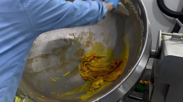 Worker scraping remaining food filling from an industrial mixing kettle after production. Product recovery process used to maximize yield, reduce food waste, and improve manufacturing efficiency in fo