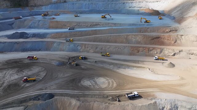 Aerial tracking shot of excavators and dump trucks loading and hauling earth in open pit quarry terraces during daytime industrial operation