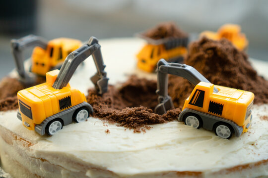 Homemade construction birthday cake with three yellow toy excavators on white frosting and brown crumbs. Playful building site dessert for kid party. Crumbled biscuit as sand and earthwork by mom