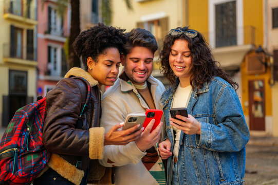 Group of young diverse friends smiling and sharing content on their mobile phones while traveling and navigating an old european city together for a social vacation
