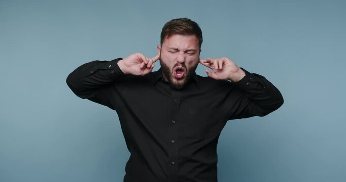 Man covers ears while making funny faces in indoor setting with blue background during day