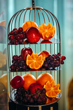 three-tier metal cage fruit stand showcasing carved oranges, red apples, and purple grapes, shot with soft natural lighting against a blurred light-green curtain background