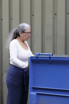 Female worker with an expression of disgust and revulsion on face, looks inside an industrial waste container on farm, belonging to integrated waste management services or industrial waste collection
