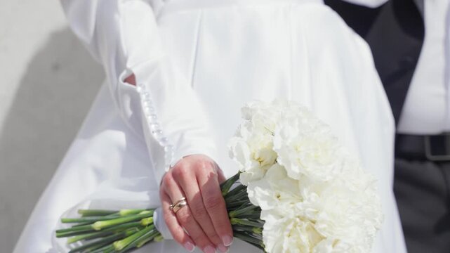 Elegant close-up shot of bride in white dress holding white carnation bouquet, with groom beside her. Bright natural lighting highlights fine details and romantic atmosphere. 198 characters.