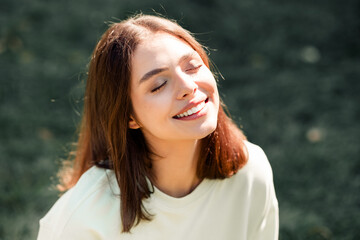 Naklejka premium Young woman smiling with closed eyes enjoying sunlight outdoors in a relaxed casual portrait