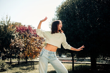 Obraz premium Happy young woman dancing in a sunlit park wearing a casual crop top and jeans, carefree summer lifestyle portrait expressing joy and movement