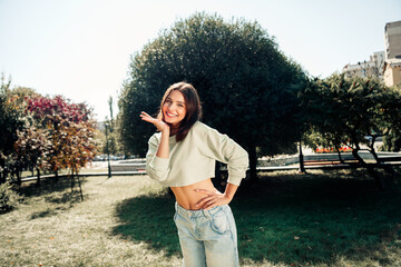 Fototapeta premium Smiling young woman in cropped sweatshirt and jeans posing playfully in a sunny city park