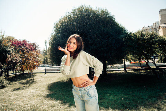 Smiling young woman in cropped sweatshirt and jeans posing playfully in a sunny city park