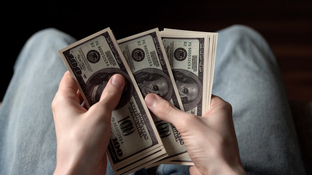 A young man is holding a stack of dollar bills in his hands, counting the money. The man appears focused on the dollar bills, revealing the intricate details of currency.