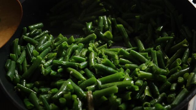 Hand with wooden spoon stirring green bean pods in pan. Preparing plant based vegetarian meal