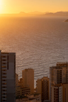 Sunset over Benidorm Spain coastline glows across Mediterranean sea, with apartment towers, resort skyline and mountains fading in warm haze