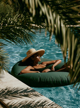 Young Woman Relaxing in Inflatable Ring Floating in Blue Swimming Pool With Straw Hat in Lush Mediterranean Summer Resort Setting
