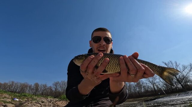 Young smiling fisherman in sunglasses holding caught European chub [Squalius cephalus] showing it to camera by river weir