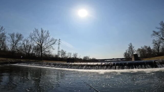 Scenic view of river weir with fast flowing water on sunny day, fishing rod tip in foreground, outdoor relaxation landscape