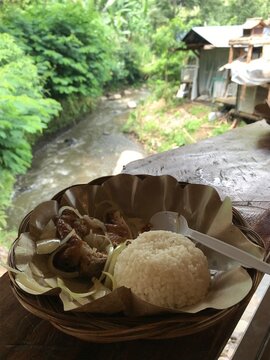 Rustic grilled chicken and rice meal overlooking a flowing river and lush tropical vegetation, ideal for travel, food, nature, and lifestyle concepts.