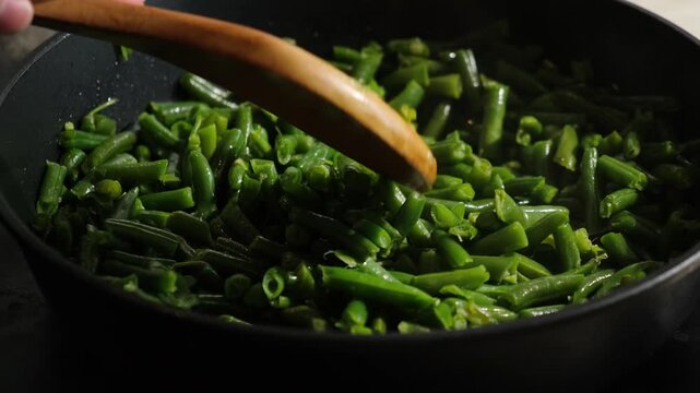 Hand with wooden spoon stirring green bean pods in frying pan