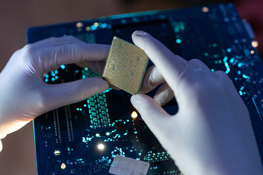 A person with white gloves holds a computer chip above a circuit board while working on tech tasks in a dim area