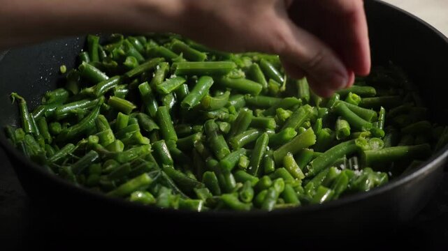 Hand sprinkling salt on green bean pods frying in a pan. Cooking plant based vegetarian meal