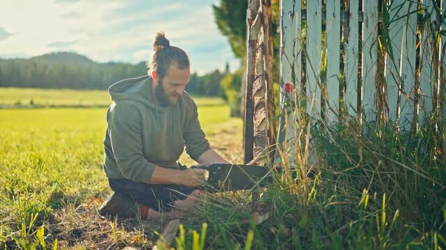 A Man is Kneeling by a Rustic Wooden Fence, Sawing Off the Excess Timber in a Grassy Field Under Daylight - Medium Shot